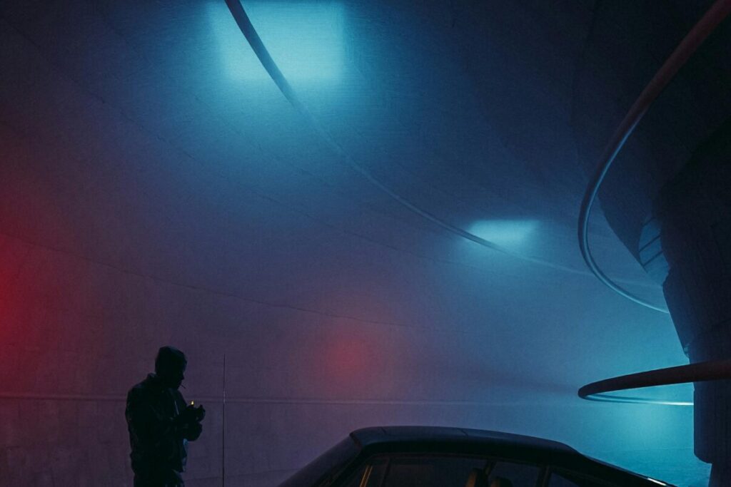 Silhouette of a person by a vintage car in a moody, neon-lit tunnel.